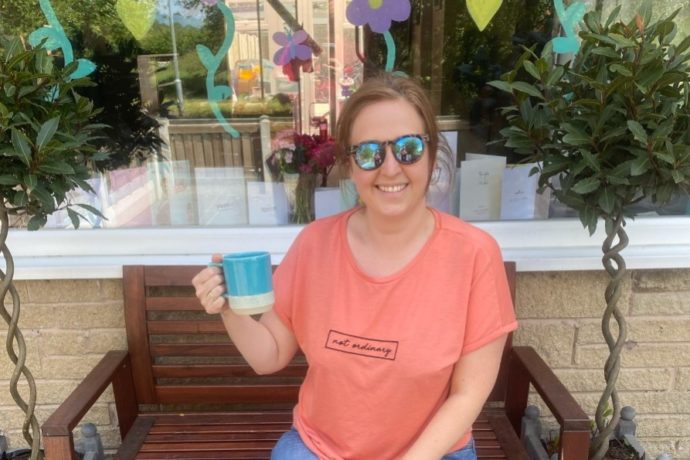 Louise sits on a bench in her garden, holding up a cup of tea to say cheers